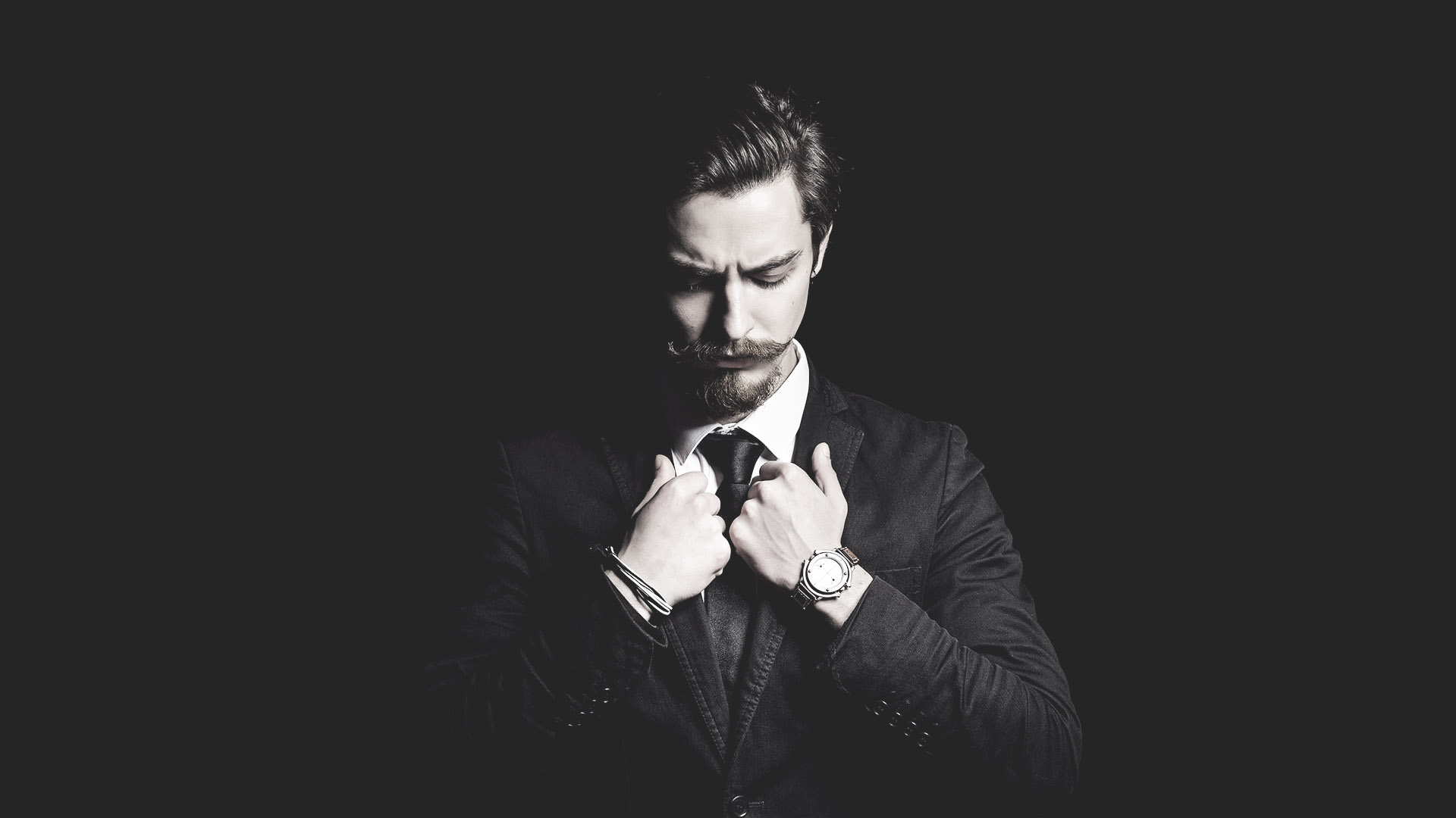 Man in a black suit adjusting his tie with a serious expression.